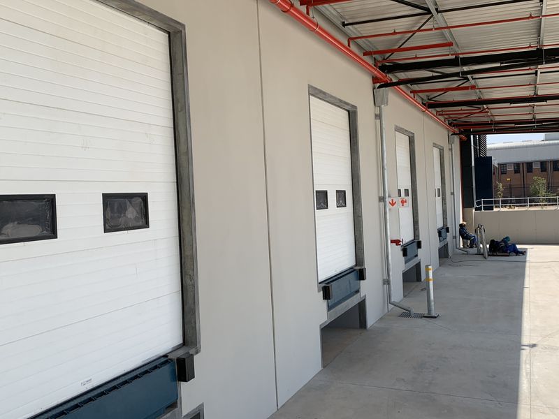 Industrial loading bay area featuring multiple roller shutter doors, dock levelers, and exposed fire suppression systems on a concrete apron.