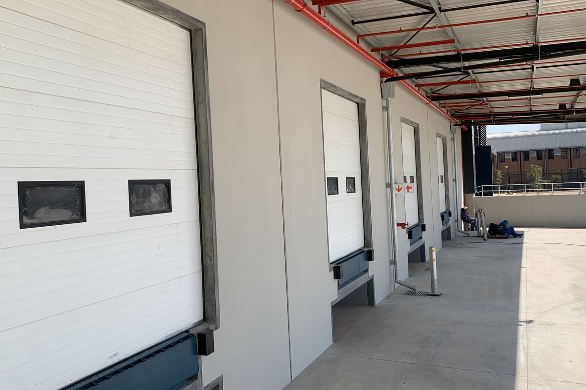 Industrial loading bay area featuring multiple roller shutter doors, dock levelers, and exposed fire suppression systems on a concrete apron.