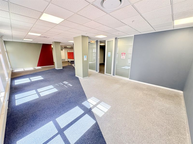 A spacious, vacant open-plan office area featuring modern glass partitions, suspended ceilings with recessed lighting, and clean carpeted flooring.