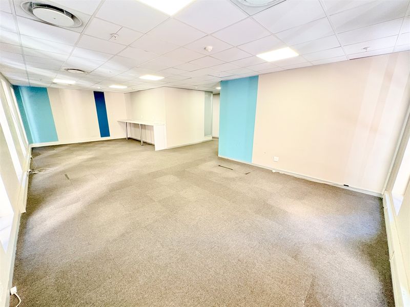 An empty, open-plan office space featuring suspended ceilings, recessed lighting, and neutral carpet flooring with blue accent wall detailing.