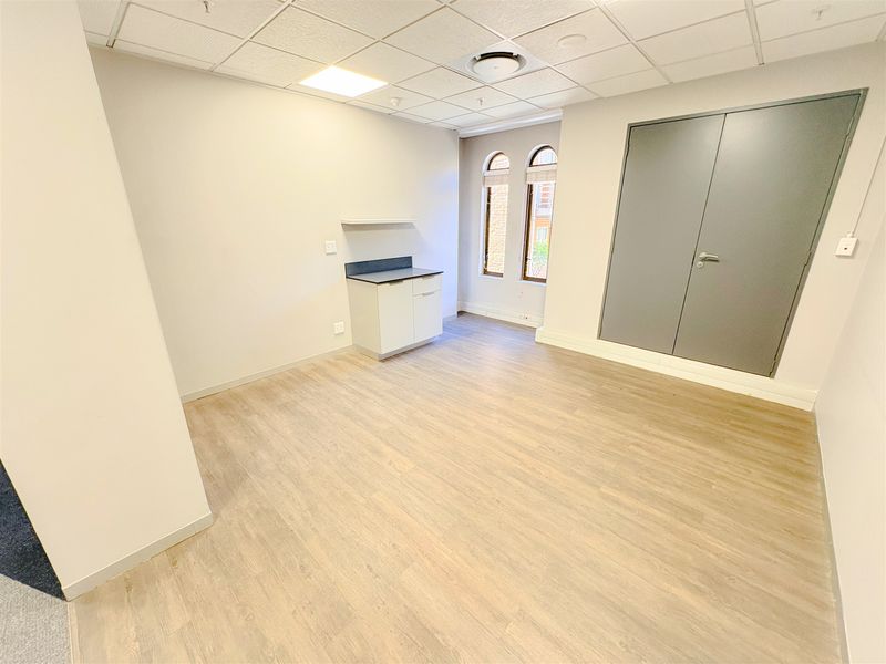 A bright, vacant office space featuring modern laminate flooring, a suspended ceiling with recessed lighting, and a compact built-in kitchenette unit.