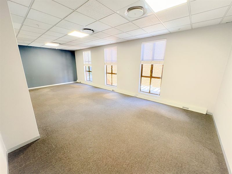 A bright, open-plan office space featuring neutral carpeting, a suspended ceiling with recessed lighting, and large windows with venetian blinds.