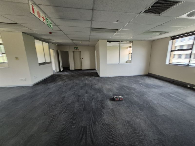 A spacious, empty open-plan office area featuring carpet tiles, a suspended ceiling, and large windows providing natural light.