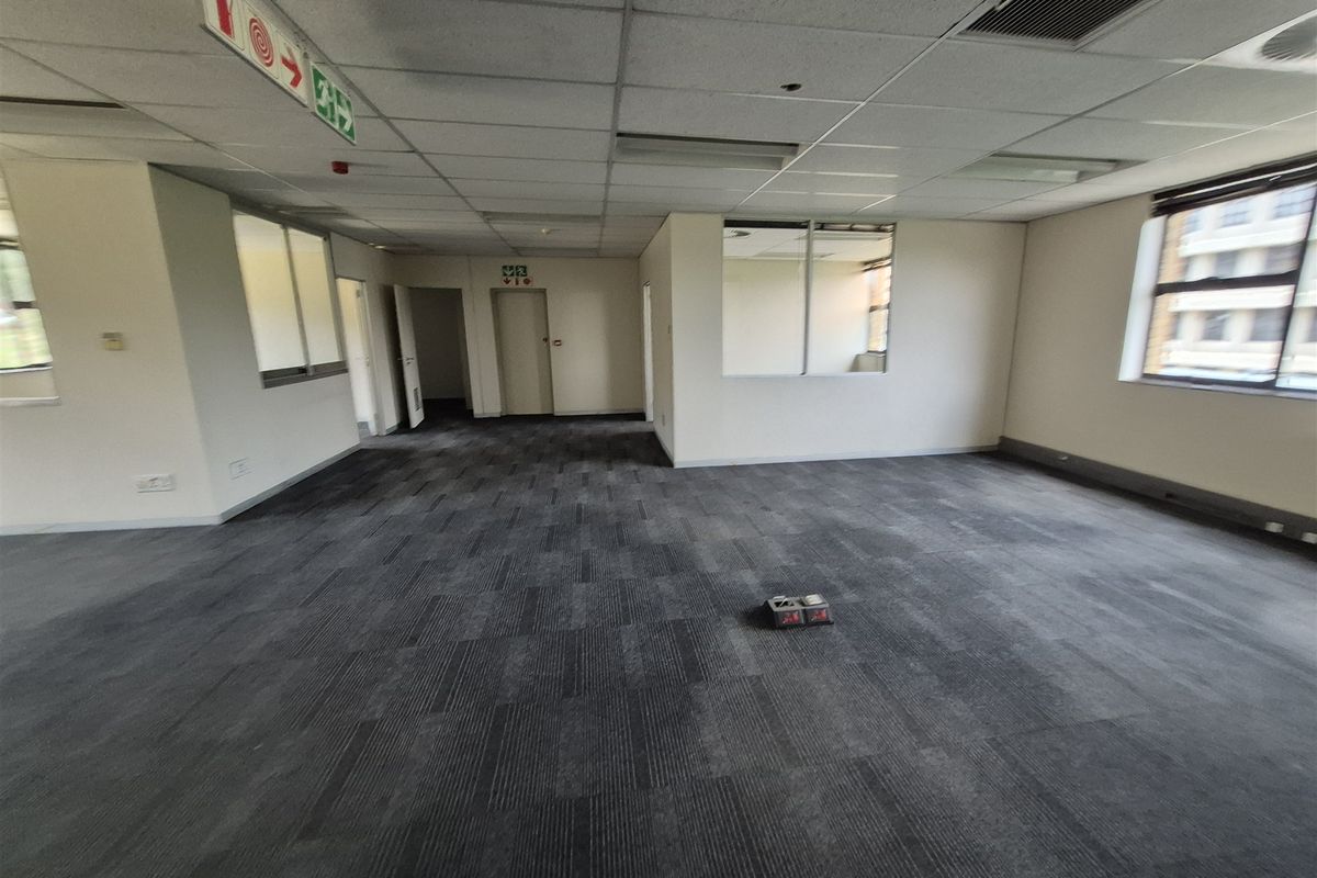 A spacious, empty open-plan office area featuring carpet tiles, a suspended ceiling, and large windows providing natural light.