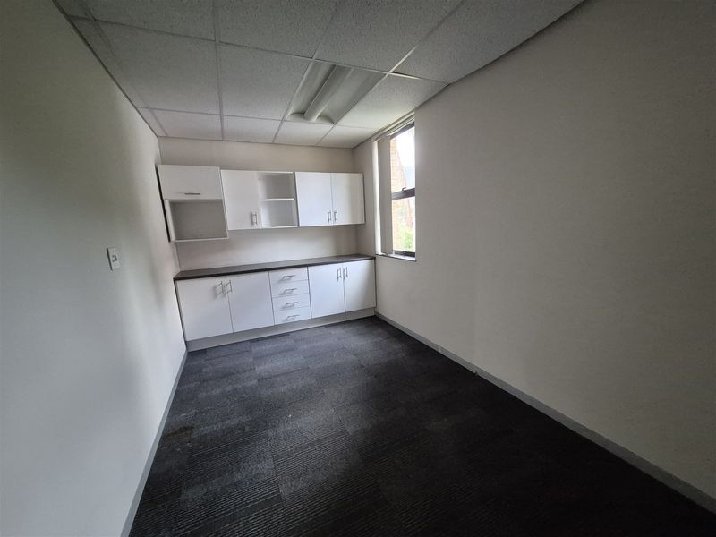 A vacant office space featuring built-in white cabinetry, dark carpet tiles, and a drop ceiling with integrated lighting.