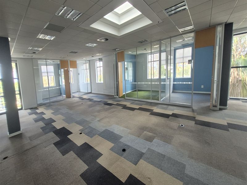 Open-plan office space featuring glass-partitioned private offices, a central skylight, and modern patterned carpet tiles.