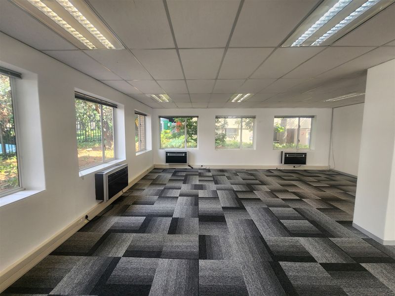 A spacious, empty office area featuring carpet tiles, suspended ceilings with recessed lighting, and multiple window-mounted air conditioning units.