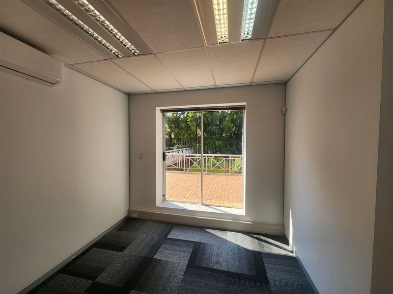A bright, empty office space featuring carpet tiles, a suspended ceiling with recessed lighting, a wall-mounted air conditioner, and a sliding door to a balcony.