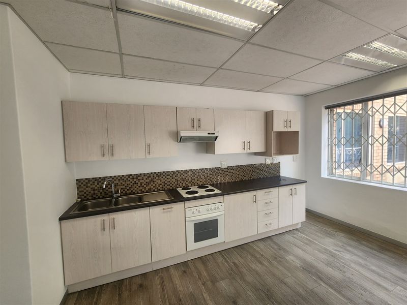 A clean, functional office kitchenette featuring light wood-grain cabinetry, dark laminate countertops, a built-in oven, and a tiled backsplash.