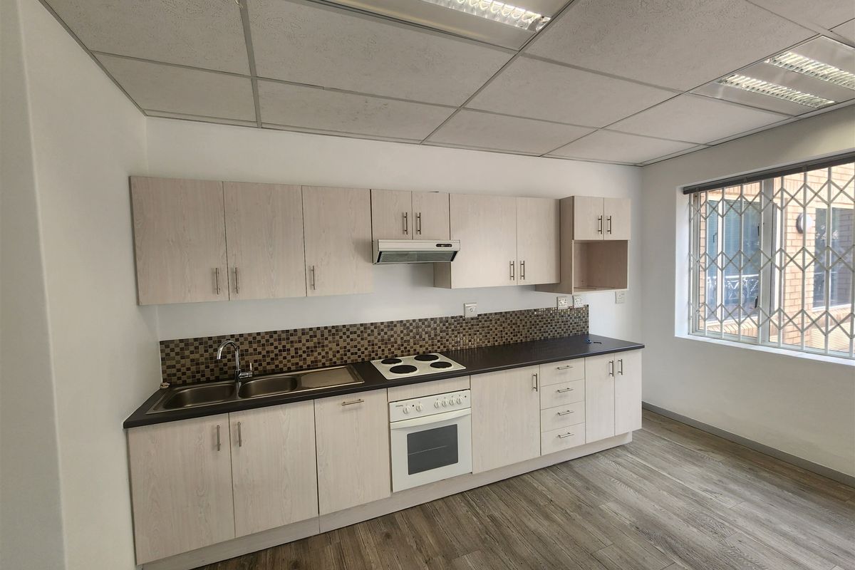 A clean, functional office kitchenette featuring light wood-grain cabinetry, dark laminate countertops, a built-in oven, and a tiled backsplash.