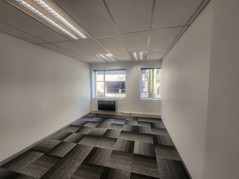 A clean, empty office space featuring neutral carpet tiles, a suspended ceiling with integrated lighting, and a window-mounted air conditioning unit.