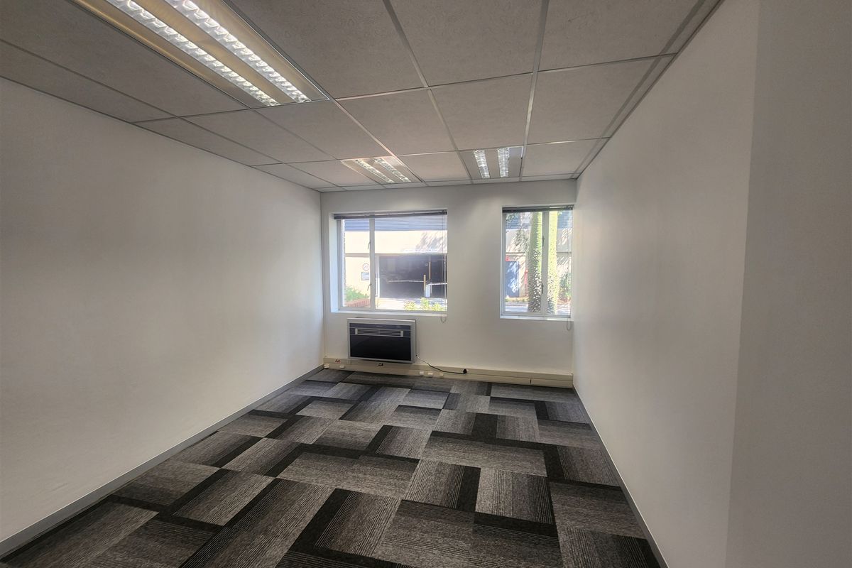 A clean, empty office space featuring neutral carpet tiles, a suspended ceiling with integrated lighting, and a window-mounted air conditioning unit.