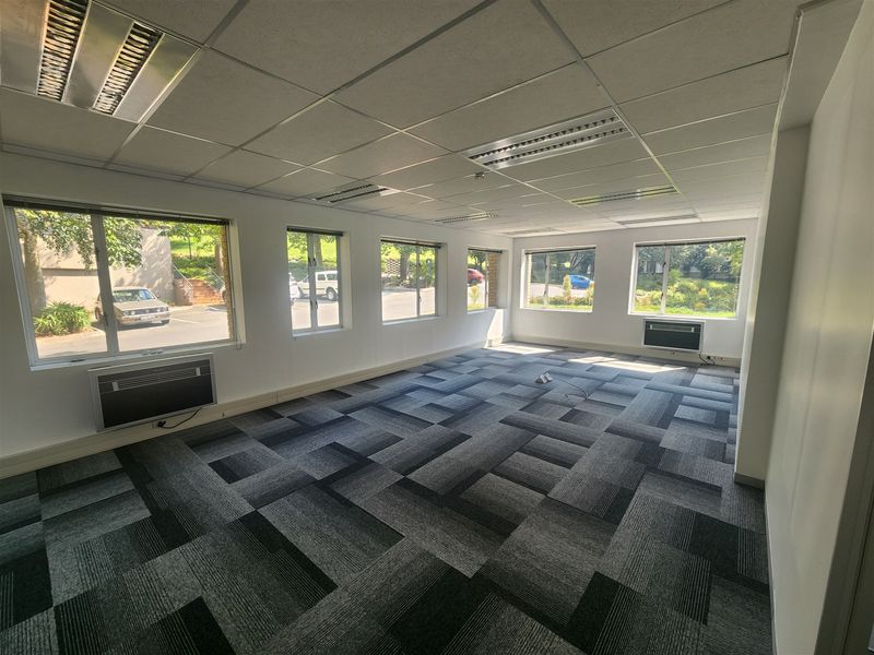 A spacious, empty office area featuring carpet tiles, suspended ceilings with recessed lighting, and multiple window-mounted air conditioning units.