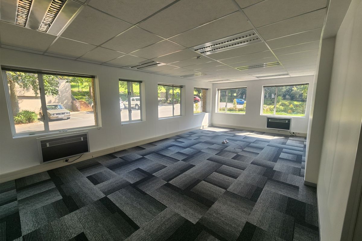 A spacious, empty office area featuring carpet tiles, suspended ceilings with recessed lighting, and multiple window-mounted air conditioning units.