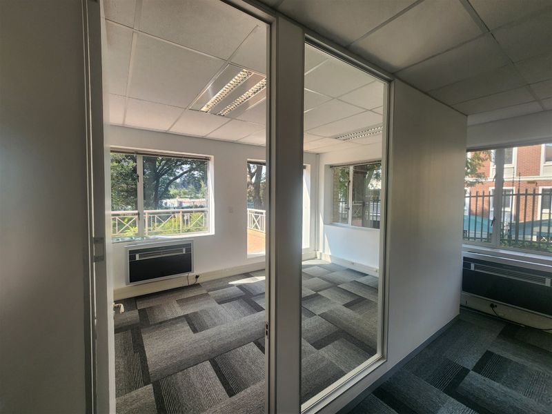A bright, vacant office space featuring glass partitions, carpet tiles, suspended ceilings with recessed lighting, and integrated window air conditioning units.