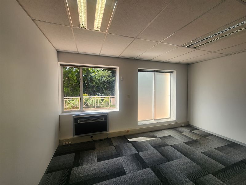 A vacant office space featuring carpet tiles, a suspended ceiling with recessed lighting, and a window-mounted air conditioning unit.