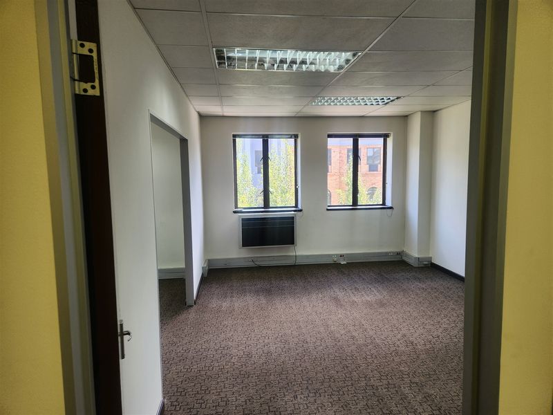 An empty office space featuring a suspended ceiling with recessed lighting, carpeted flooring, and a wall-mounted air conditioning unit.