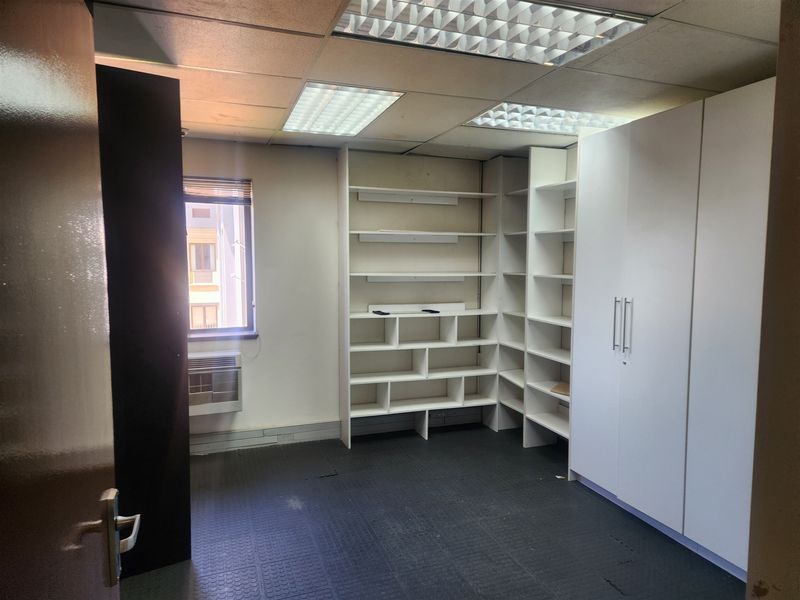 A functional office space featuring built-in shelving, ample storage cabinetry, suspended ceiling tiles, and a window-mounted air conditioning unit.