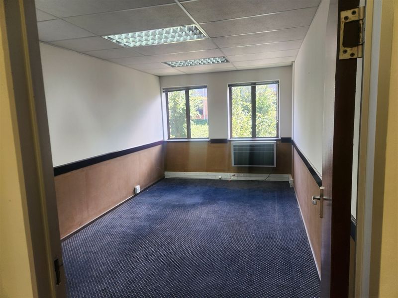An empty office space featuring a suspended ceiling, recessed lighting, a window-mounted air conditioning unit, and carpeted flooring.