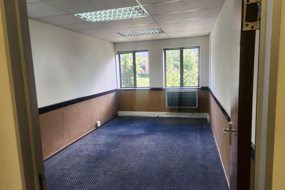An empty office space featuring a suspended ceiling, recessed lighting, a window-mounted air conditioning unit, and carpeted flooring.