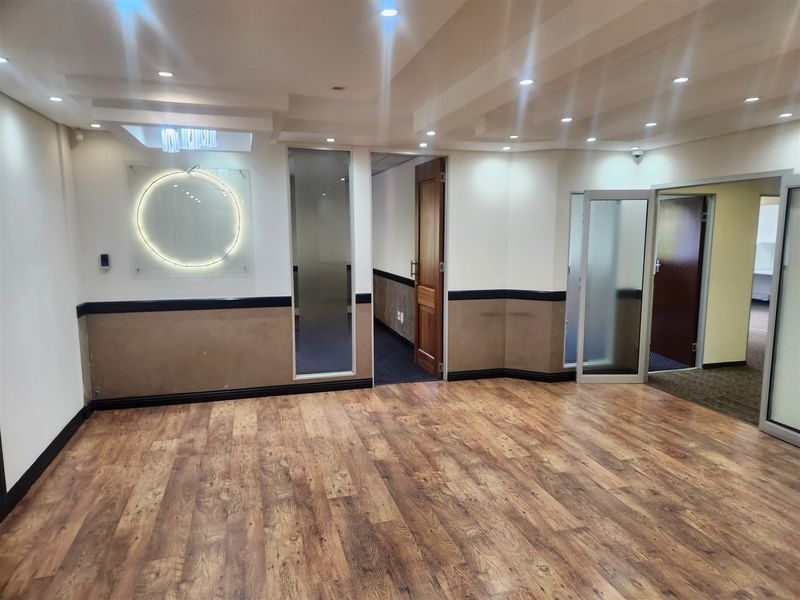 A spacious, empty office area featuring wood-look laminate flooring, recessed ceiling lighting, and multiple glass-paneled partition doors.