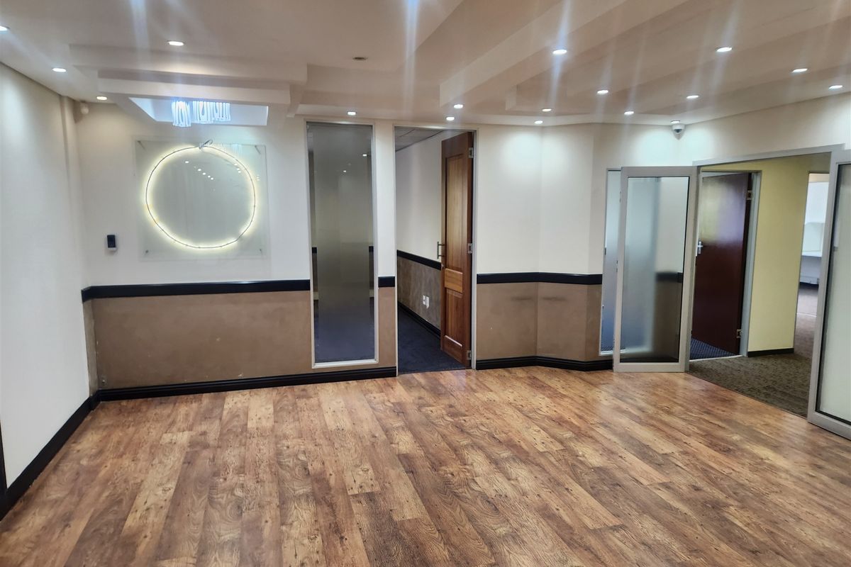 A spacious, empty office area featuring wood-look laminate flooring, recessed ceiling lighting, and multiple glass-paneled partition doors.