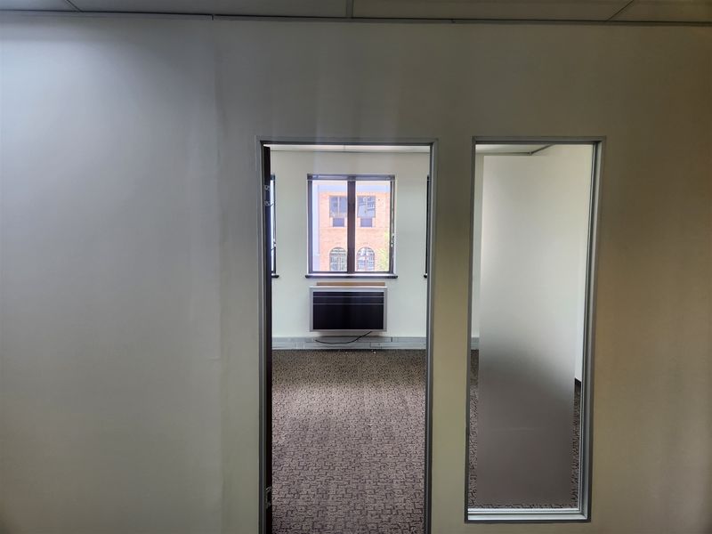 An empty office space featuring carpeted flooring, a suspended ceiling, and a window-mounted air conditioning unit with a frosted glass partition.