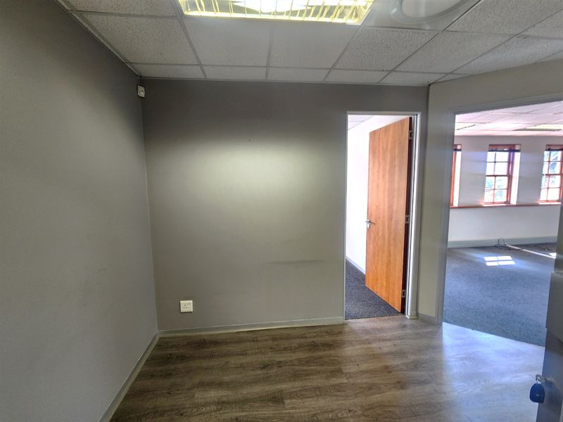 An empty office space featuring laminate flooring, a suspended ceiling with recessed lighting, and a wooden door leading to an adjacent room.