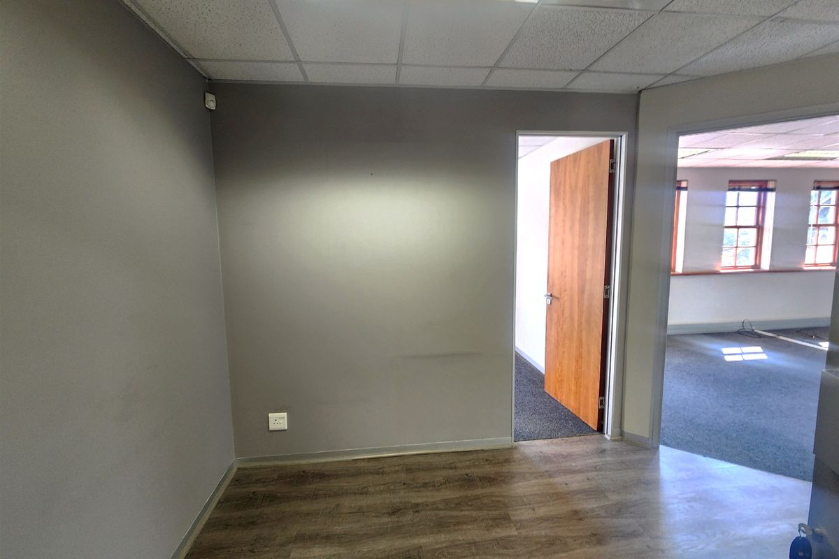 An empty office space featuring laminate flooring, a suspended ceiling with recessed lighting, and a wooden door leading to an adjacent room.