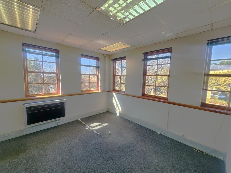 A bright, empty office space featuring multiple sash windows, carpeted flooring, a suspended ceiling with recessed lighting, and a wall-mounted air conditioning unit.
