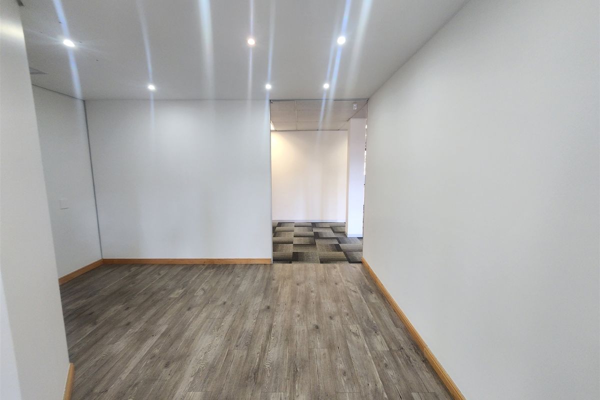 A bright, vacant office space featuring modern wood-look laminate flooring, recessed downlighting, and a transition to a carpeted area.