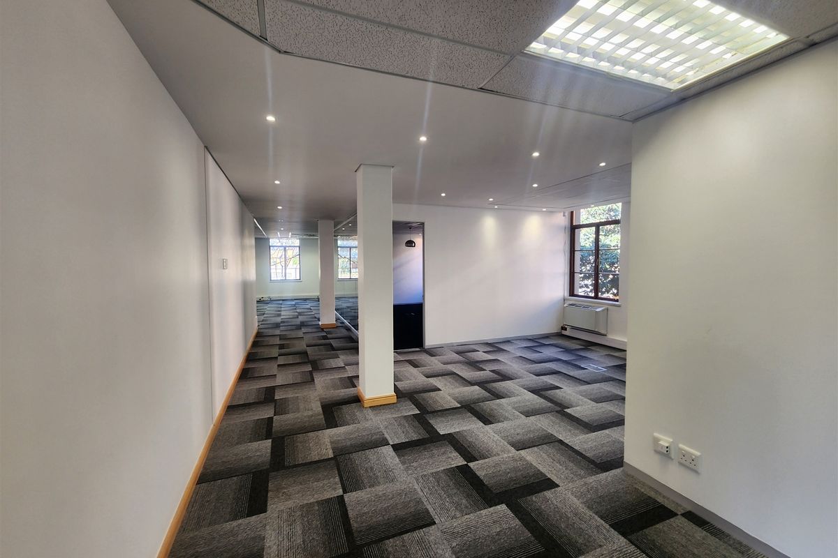A spacious, open-plan office area featuring modern carpet tiles, recessed lighting, suspended ceilings, and large windows providing natural light.
