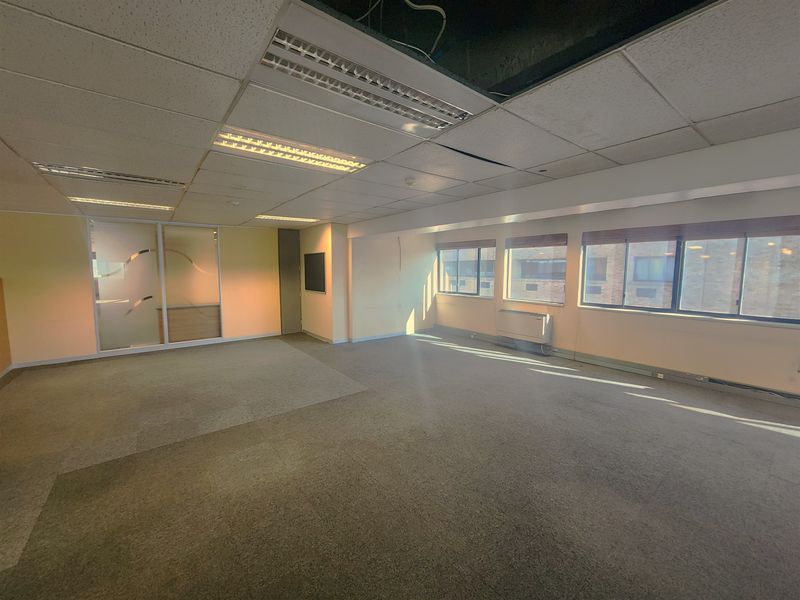 An empty, open-plan office space featuring a suspended ceiling, carpeted flooring, and large windows providing natural light.