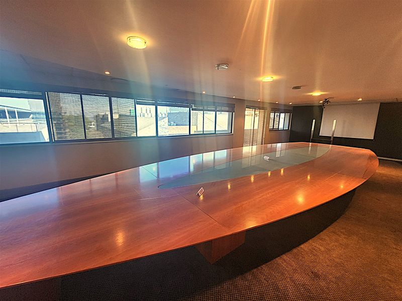 A spacious office boardroom featuring a large central conference table, carpeted flooring, recessed lighting, and a wall-mounted projection screen.