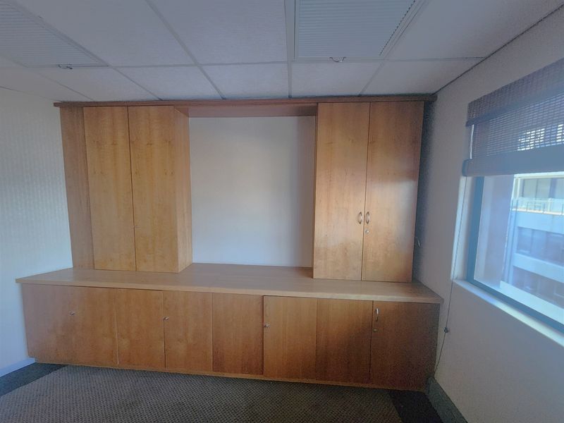 An empty office space featuring extensive built-in wooden cabinetry, a suspended ceiling, and carpeted flooring with natural light from the side window.