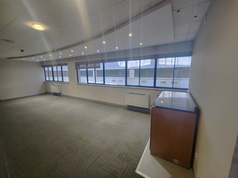 A spacious, empty office area featuring carpet tiles, suspended ceilings with recessed lighting, and large windows with built-in HVAC units.