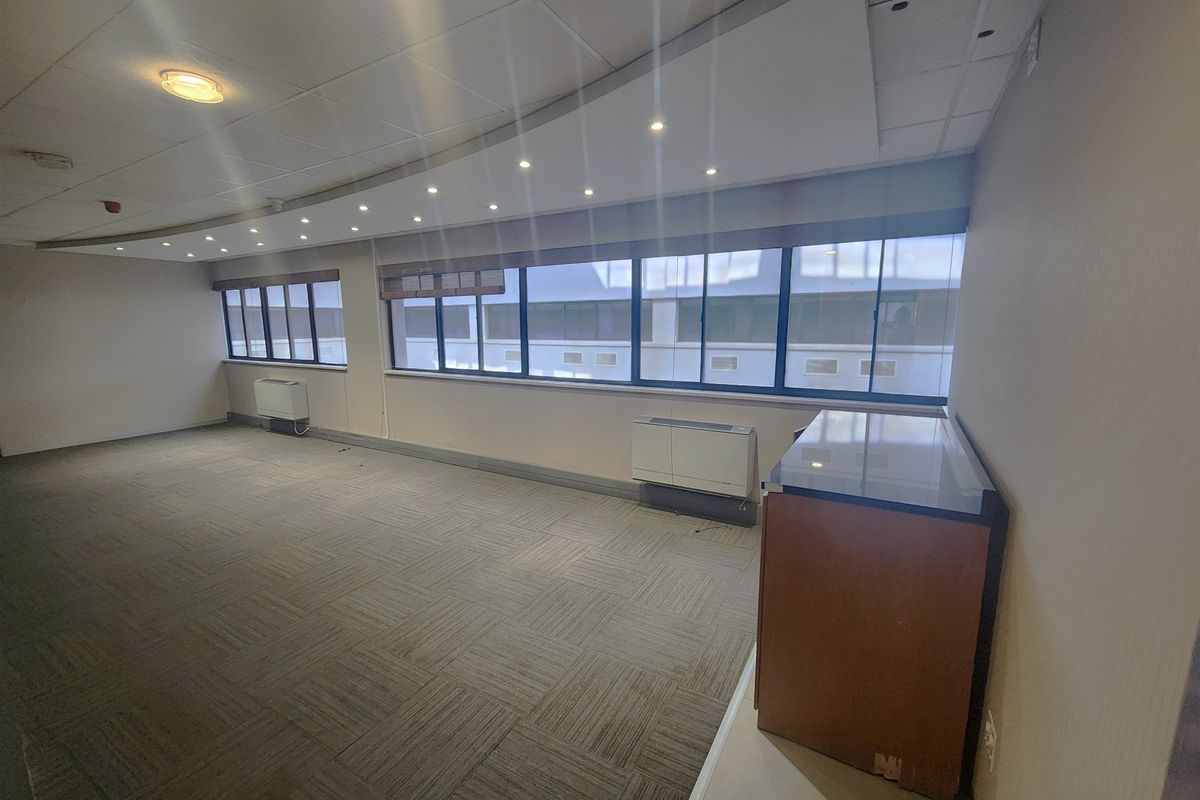 A spacious, empty office area featuring carpet tiles, suspended ceilings with recessed lighting, and large windows with built-in HVAC units.