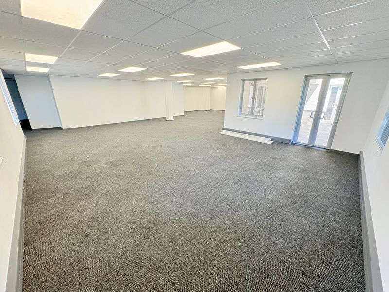 A spacious, open-plan office area featuring neutral carpet tiles, a suspended ceiling with recessed lighting, and access to an exterior balcony through glass doors.