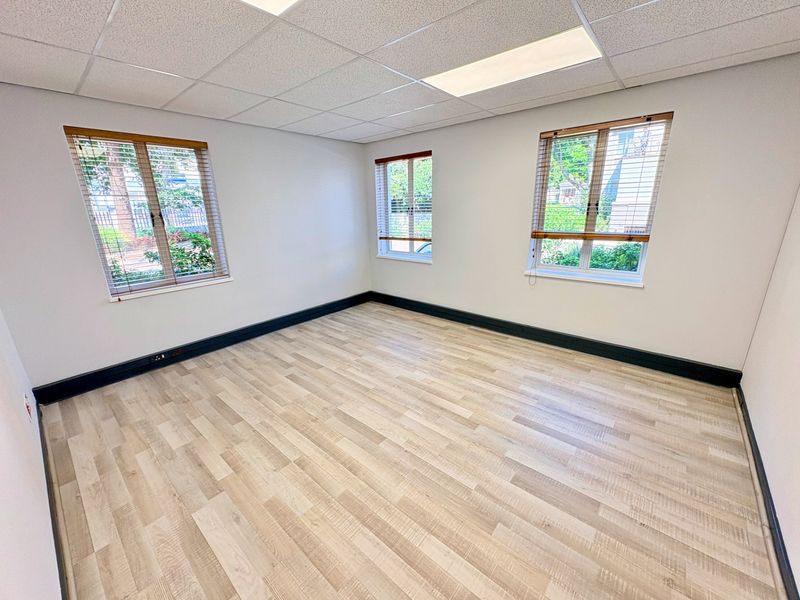 A bright, empty office space featuring modern laminate flooring, a suspended ceiling with recessed lighting, and large windows with fitted venetian blinds.