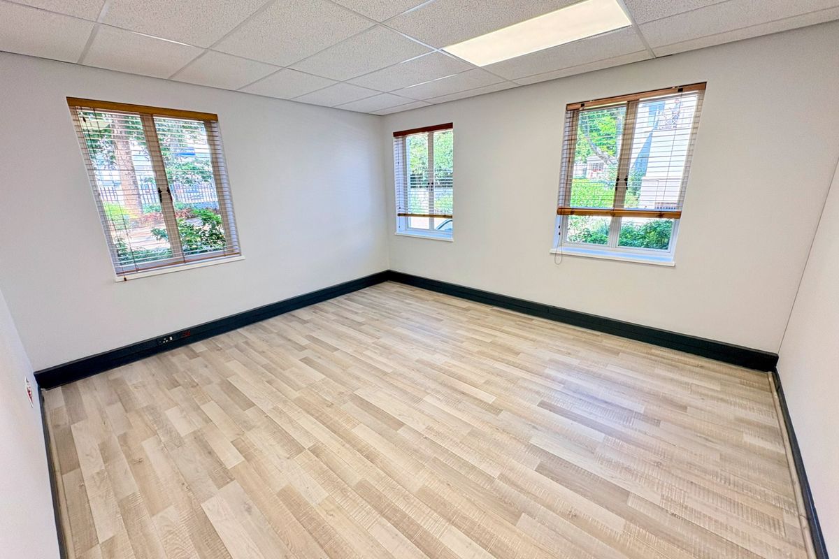 A bright, empty office space featuring modern laminate flooring, a suspended ceiling with recessed lighting, and large windows with fitted venetian blinds.
