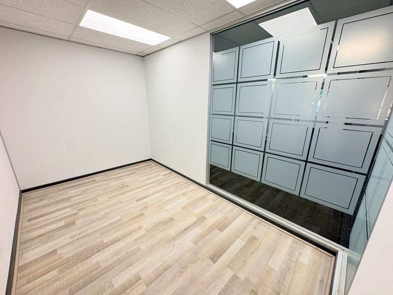 A modern, empty office space featuring light wood-look laminate flooring, a suspended ceiling with recessed lighting, and a contemporary glass partition wall with privacy film.