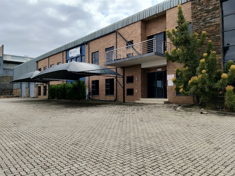 Front exterior view of an industrial property featuring a brick facade, paved driveway, shade cloth carport, and a balcony with metal railing.