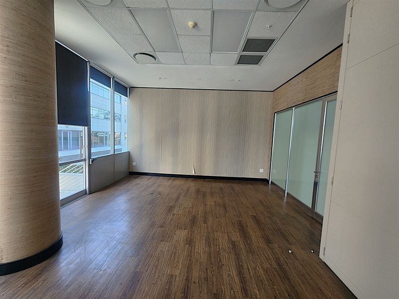 A vacant office space featuring wood-look flooring, a glass partition wall, large windows with roller blinds, and a suspended ceiling with integrated lighting.