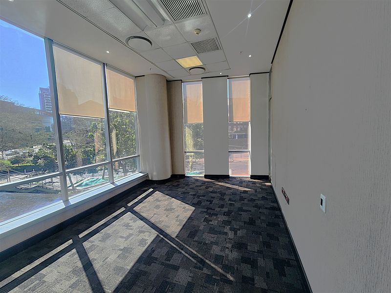 A bright, vacant office space featuring large floor-to-ceiling windows, modern carpet tiles, and a suspended ceiling with integrated lighting and air conditioning.