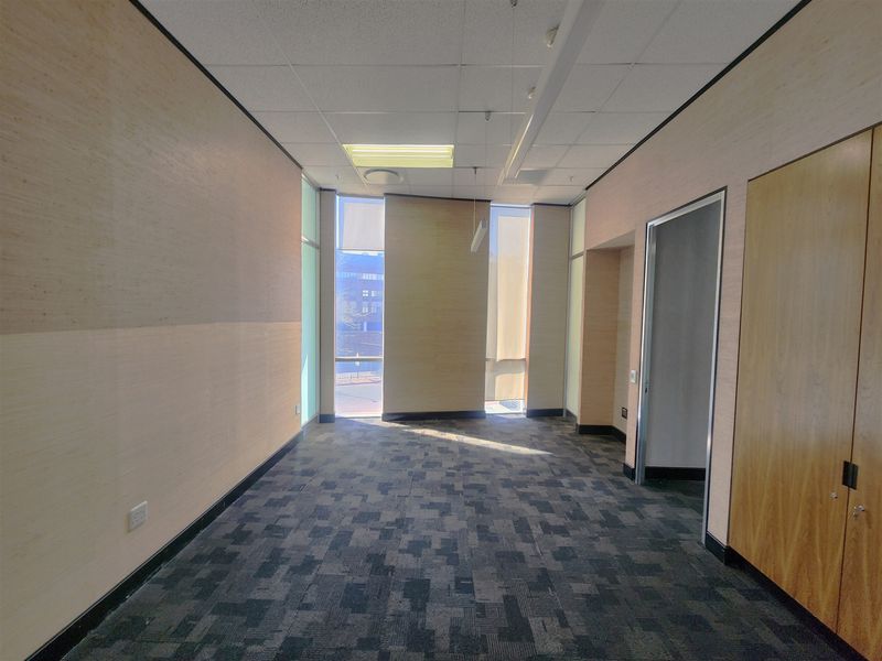 A bright, empty office space featuring carpet tiles, suspended ceilings with recessed lighting, and built-in wooden storage cabinetry.