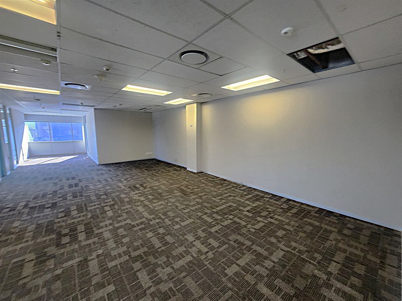 An empty, open-plan office space featuring carpeted flooring, a suspended ceiling with recessed lighting, and large windows providing natural light.