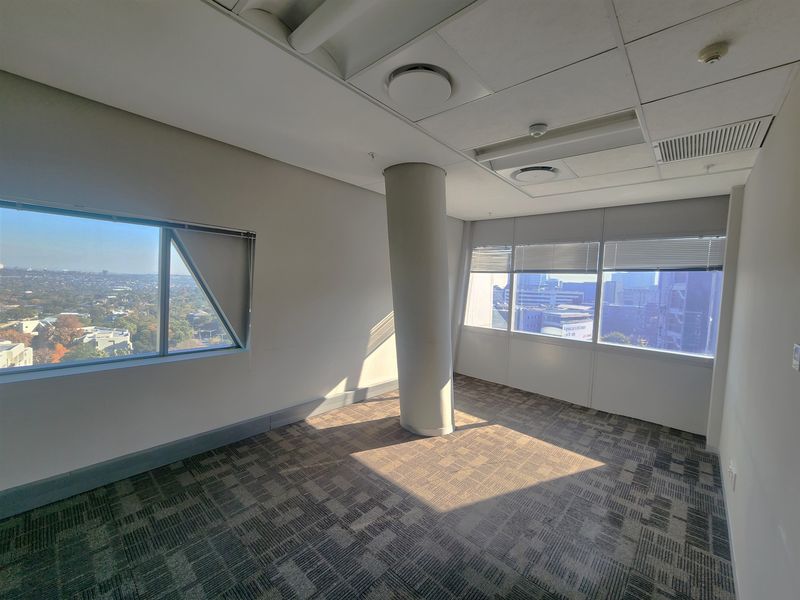 An empty, sunlit office space featuring carpet tiles, a central structural column, and large windows offering expansive city views.