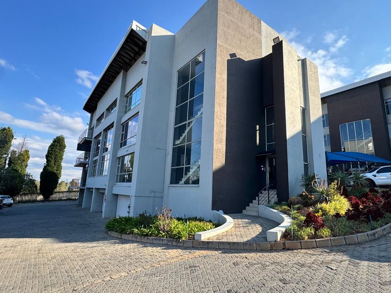Modern multi-story office building featuring large glass windows, a paved driveway, and well-maintained landscaped garden beds at the entrance.