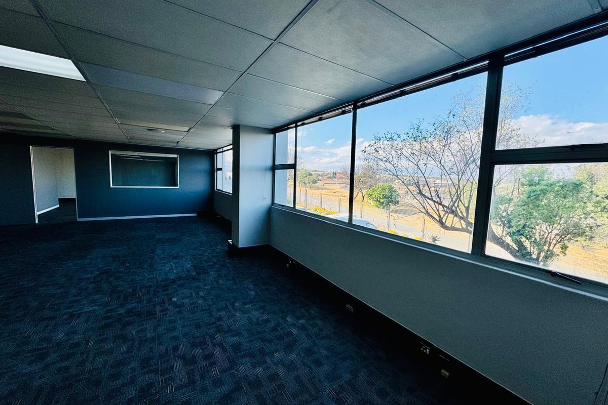 A spacious, empty office area featuring large windows with natural light, carpeted flooring, and a suspended ceiling with recessed lighting.