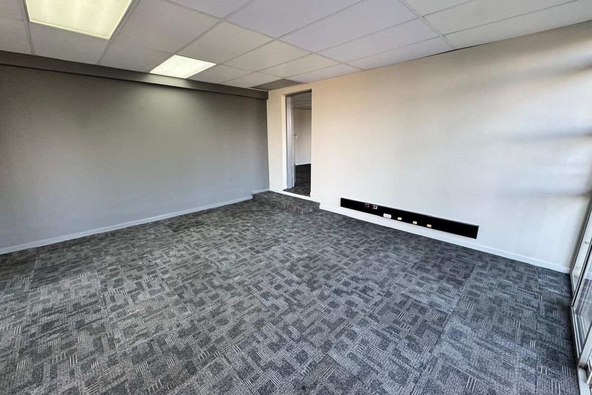 A clean, vacant office space featuring modern carpet tiles, a suspended ceiling with recessed lighting, and integrated electrical trunking along the wall.